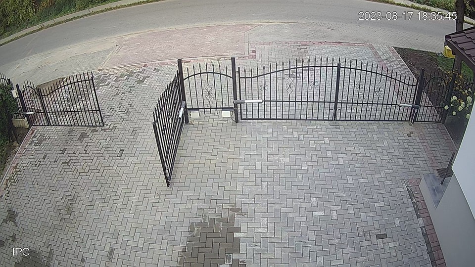 Zdalny widok na posesję z bramą automatyczną i furtką. Kostka brukowa na podjeździe, widoczne siłowniki bramy. Ujęcie z kamery monitoringu.