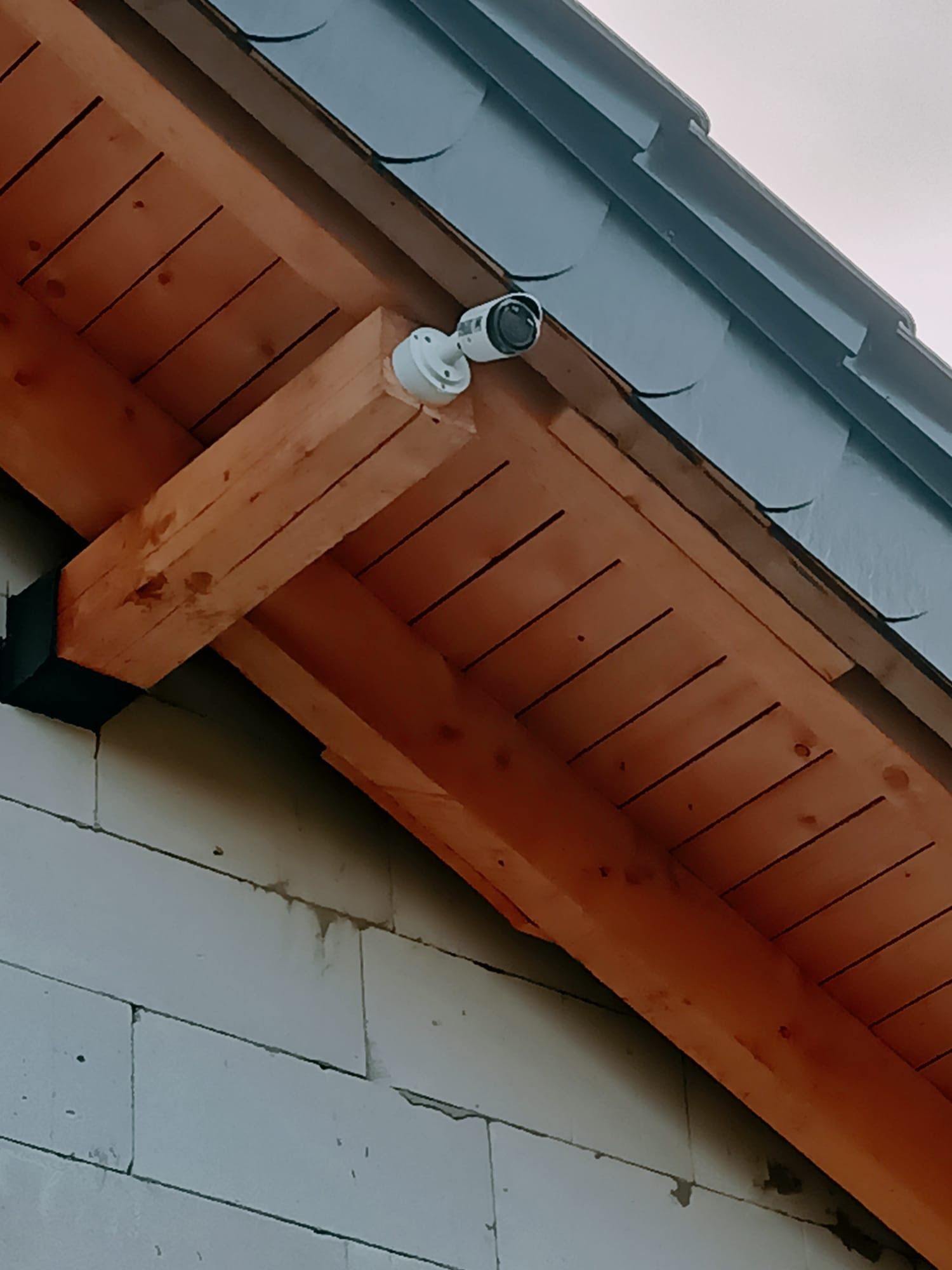 Kamera monitoringu zamontowana na drewnianej konstrukcji pod dachem z szarej dachówki, widoczna z dołu, na tle ściany z białych bloczków.