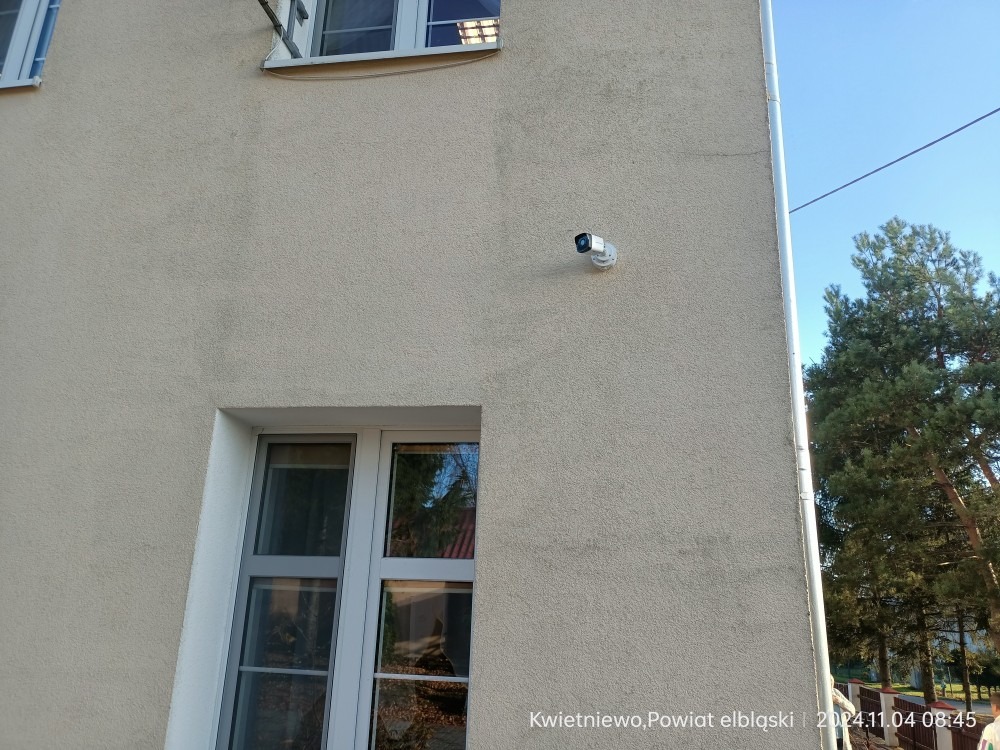 Kamera monitoringu zamontowana na elewacji budynku w pobliżu okien i rynny, z widocznym fragmentem drzewa i niebem w tle. Ujęcie z dołu.