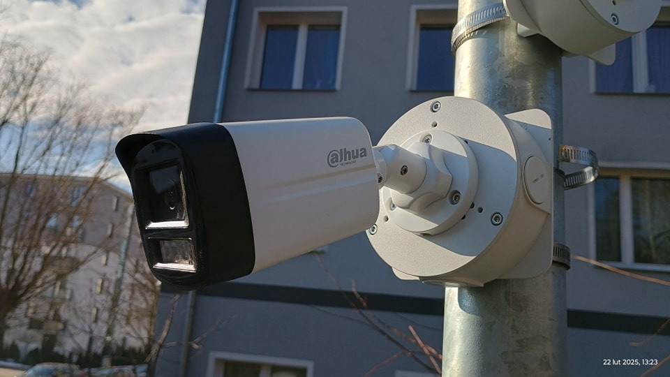 Kamera monitoringu Dahua na metalowym słupie, zamontowana na tle budynku z oknami. Widoczne elementy montażowe i przewody. Słoneczny dzień.