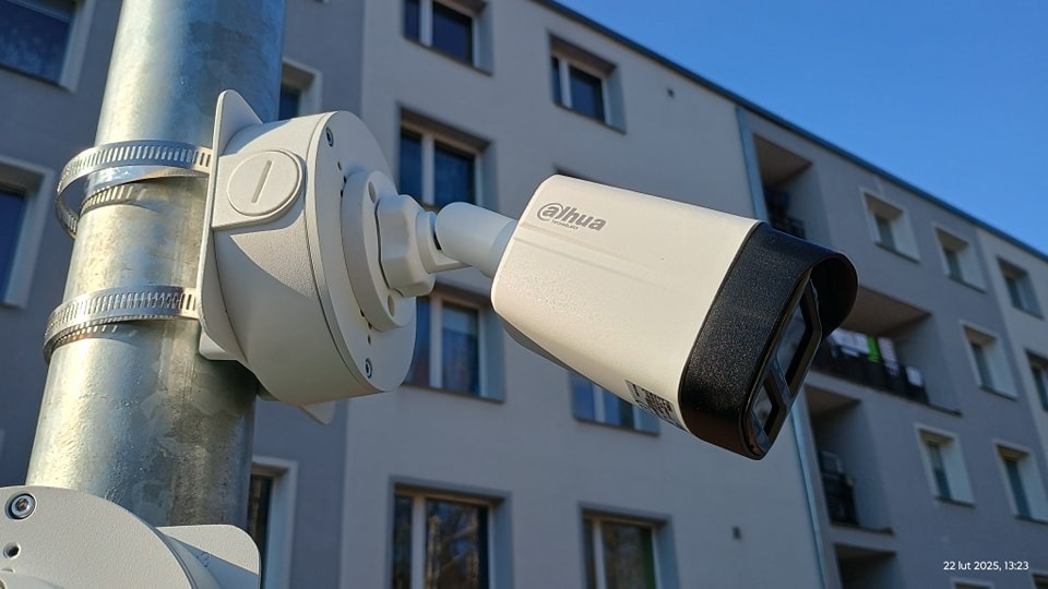 Kamera monitoringu Dahua zamocowana na słupie, z budynkiem mieszkalnym w tle. Widoczne metalowe obejmy mocujące i fragment kolejnej kamery poniżej.