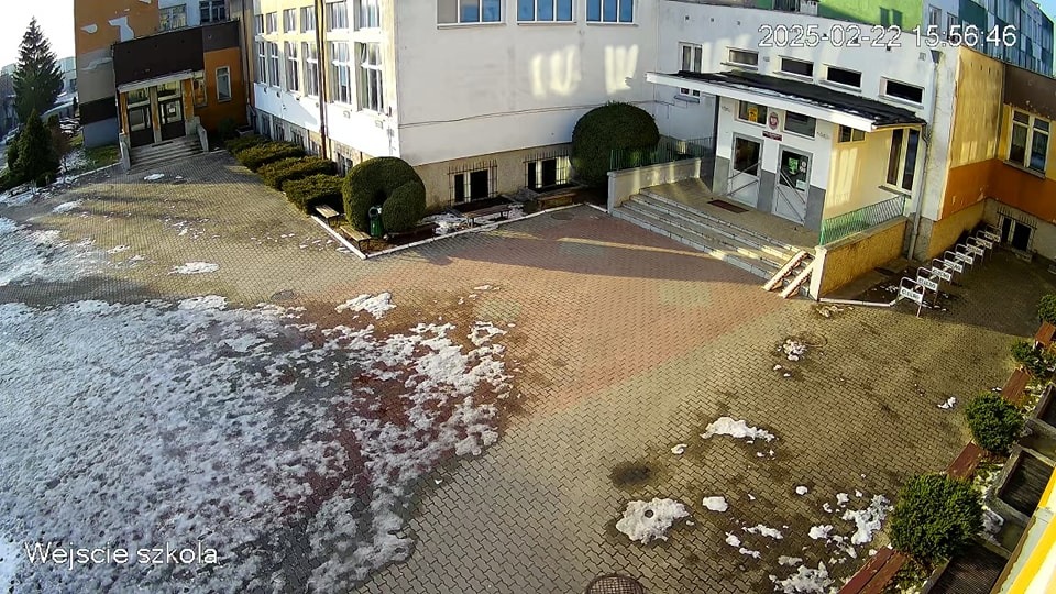 Szkolne podwórko z resztkami śniegu, widok z kamery monitoringu. Budynek szkoły w tle, kostka brukowa. Data i godzina nagrania w prawym górnym rogu.