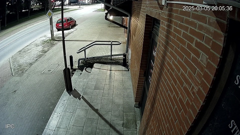 Zewnętrzna kamera monitoringu nocą, widok na ulicę i czerwony samochód. Ciemna ceglana ściana budynku po prawej. Datownik kamery w prawym górnym rogu.