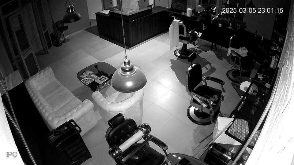 Czarno-biały obraz z kamery monitoringu ukazuje wnętrze zakładu fryzjerskiego z fotelami, kanapą w kształcie dłoni i lampami wiszącymi. Widoczny datownik kamery.