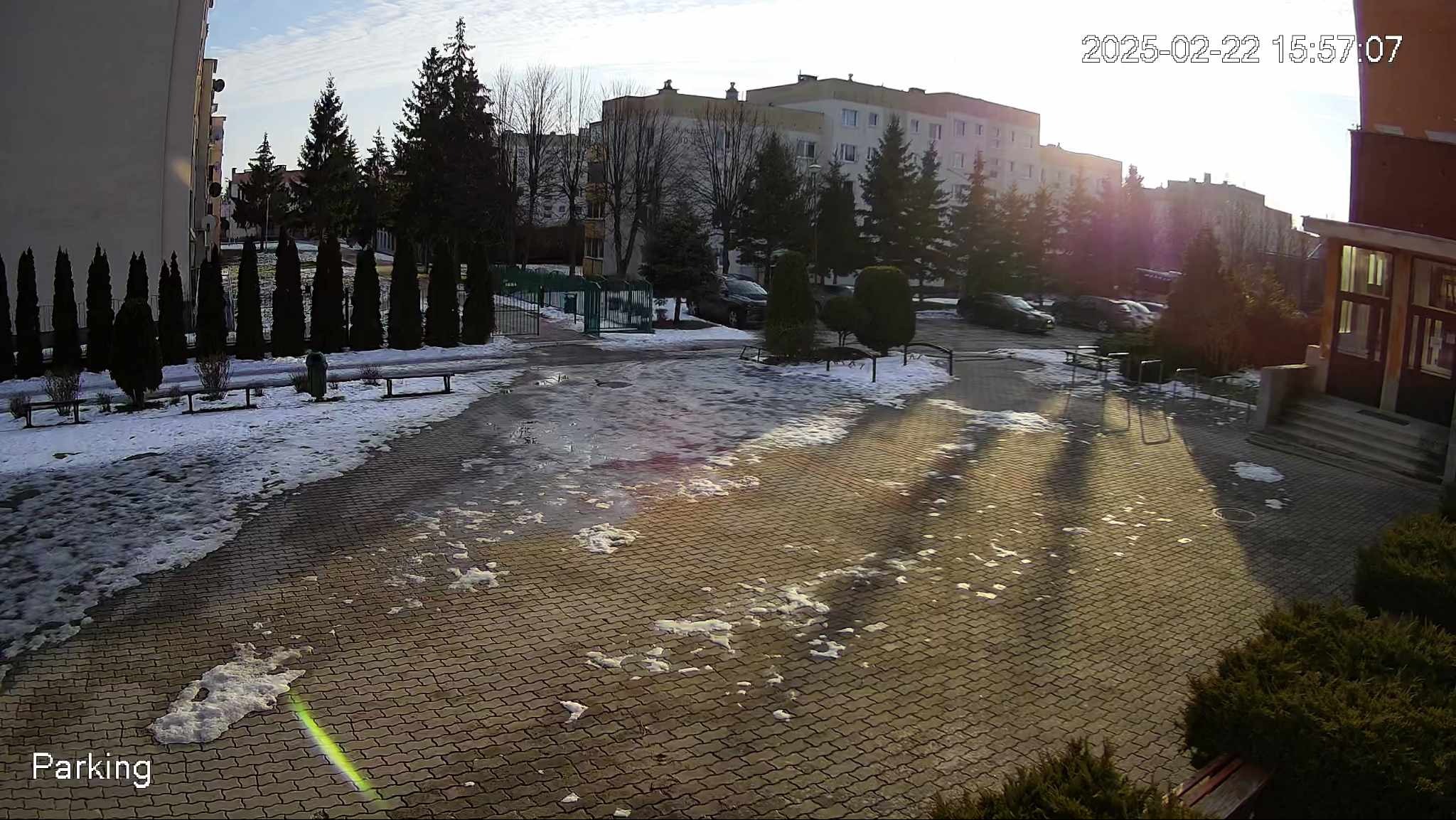 Kamera monitoringu uchwyciła widok na osiedlowy parking w zimowy dzień. Na pierwszym planie kostka brukowa z resztkami śniegu, w tle bloki i zaparkowane samochody.