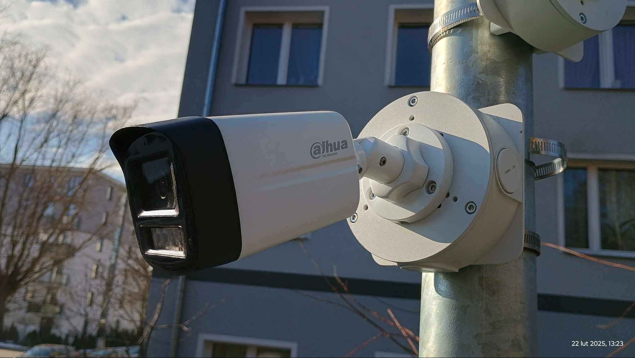 Kamera monitoringu Dahua na metalowym słupie, z budynkiem w tle. Widoczne detale montażu i obudowy kamery. Ujęcie z dołu, podkreślające wysokość instalacji.