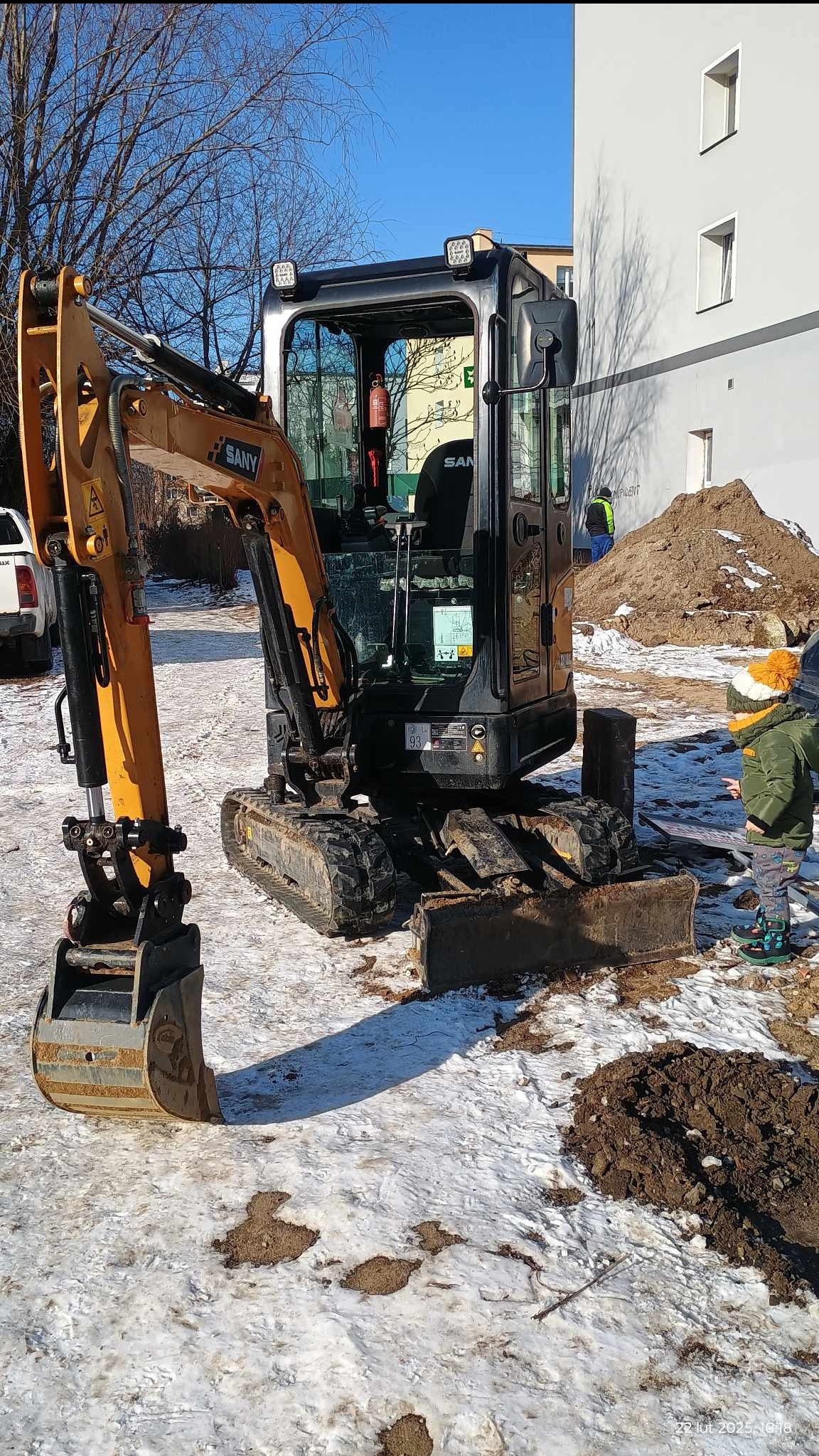 Kompaktowa koparka SANY z łyżką na śniegu, obok dziecko w zimowej kurtce i czapce z pomponem. W tle budynek i pracownik w odblaskowej kamizelce.
