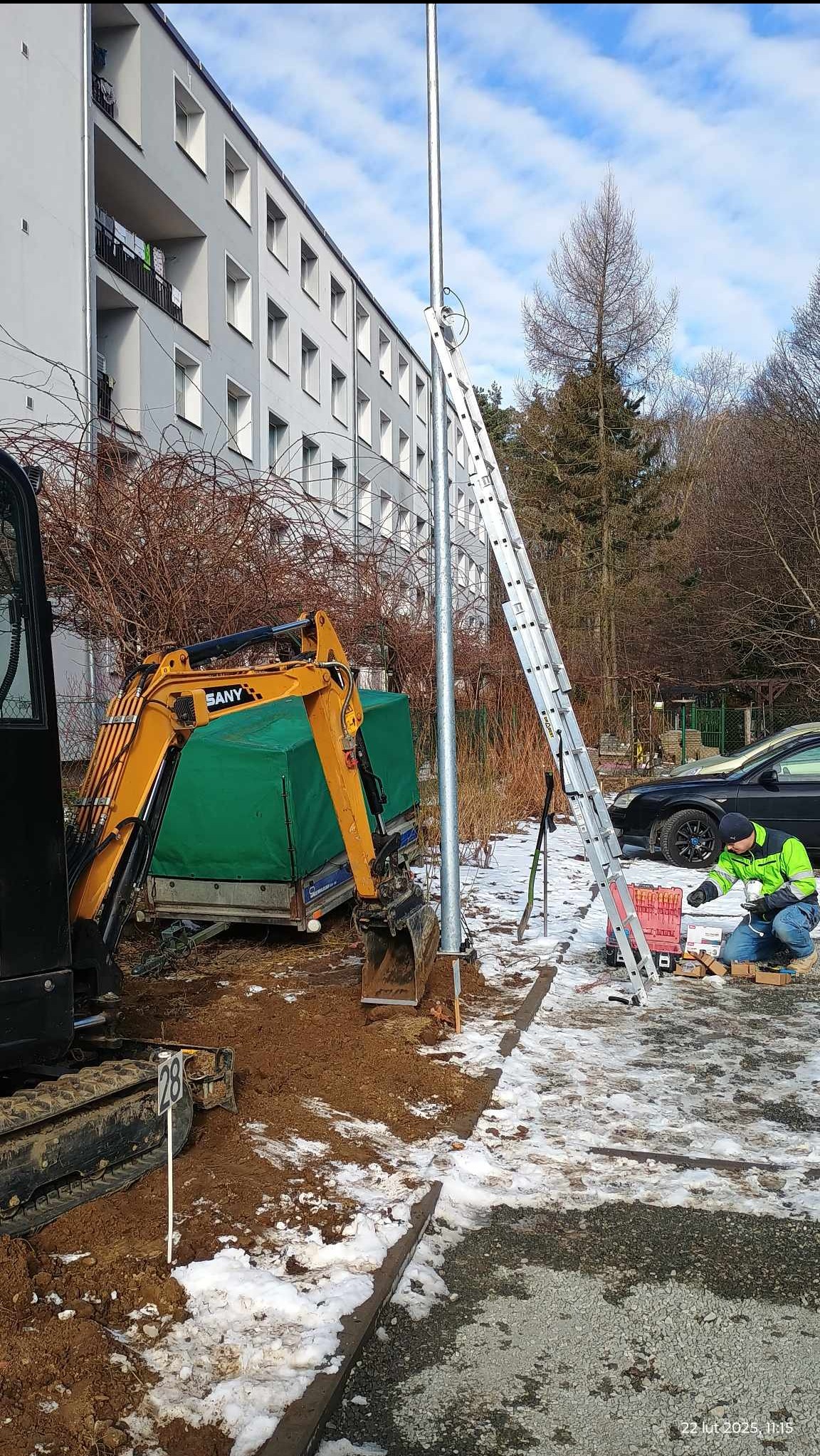 Montaż słupa oświetleniowego: koparka SANY, drabina oparta o słup, pracownik w odblaskowej kurtce przygotowuje elementy montażowe. Prace ziemne w toku.