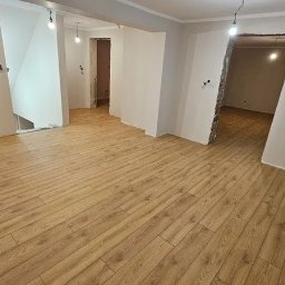 Mistrzowie - Jasne panele podłogowe w przestronnym, odnawianym wnętrzu z widocznymi przejściami do innych pomieszczeń i fragmentem niebieskiej ściany w tle.