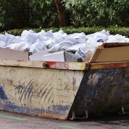 Mistrzowie - Pełny, otwarty kontener na gruz z widocznymi workami i elementami wyposażenia wnętrz na ulicy, z zieloną linią. Kontener w kolorach żółtym, czerwonym i niebieskim.