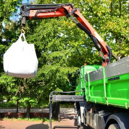 Mistrzowie - Żuraw samochodowy podnosi biały worek typu Big Bag na tle zieleni. Widoczny fragment skrzyni ładunkowej. Słoneczny dzień.