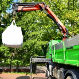 Żuraw samochodowy podnosi biały worek typu Big Bag na tle zieleni. Widoczny fragment skrzyni ładunkowej. Słoneczny dzień.