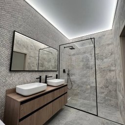Mistrzowie - Nowoczesna łazienka z szarą mozaiką, drewnianą szafką pod umywalki i kabiną prysznicową typu walk-in z czarnymi ramami, oświetlona taśmą LED.