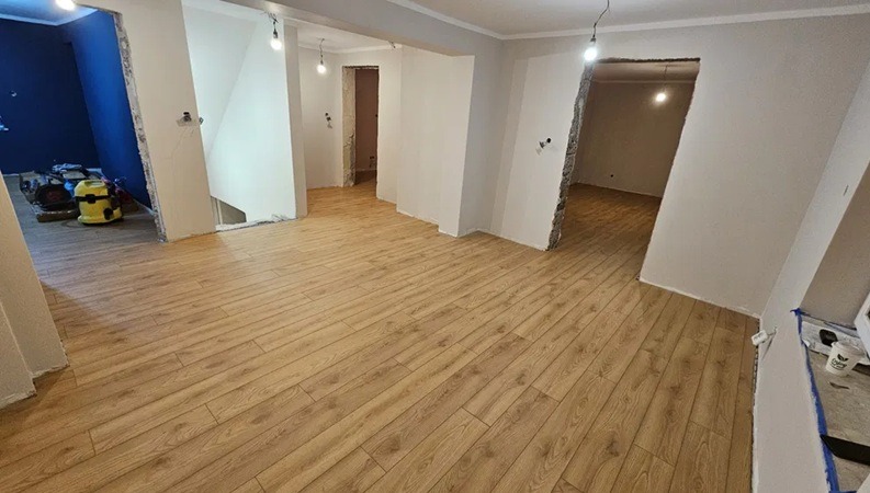 Jasne panele podłogowe w przestronnym, odnawianym wnętrzu z widocznymi przejściami do innych pomieszczeń i fragmentem niebieskiej ściany w tle.