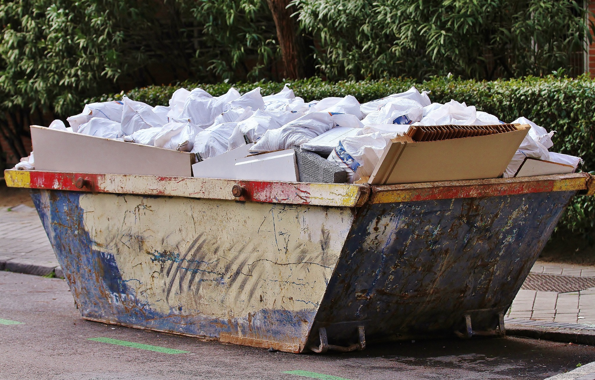 Pełny, otwarty kontener na gruz z widocznymi workami i elementami wyposażenia wnętrz na ulicy, z zieloną linią. Kontener w kolorach żółtym, czerwonym i niebieskim.
