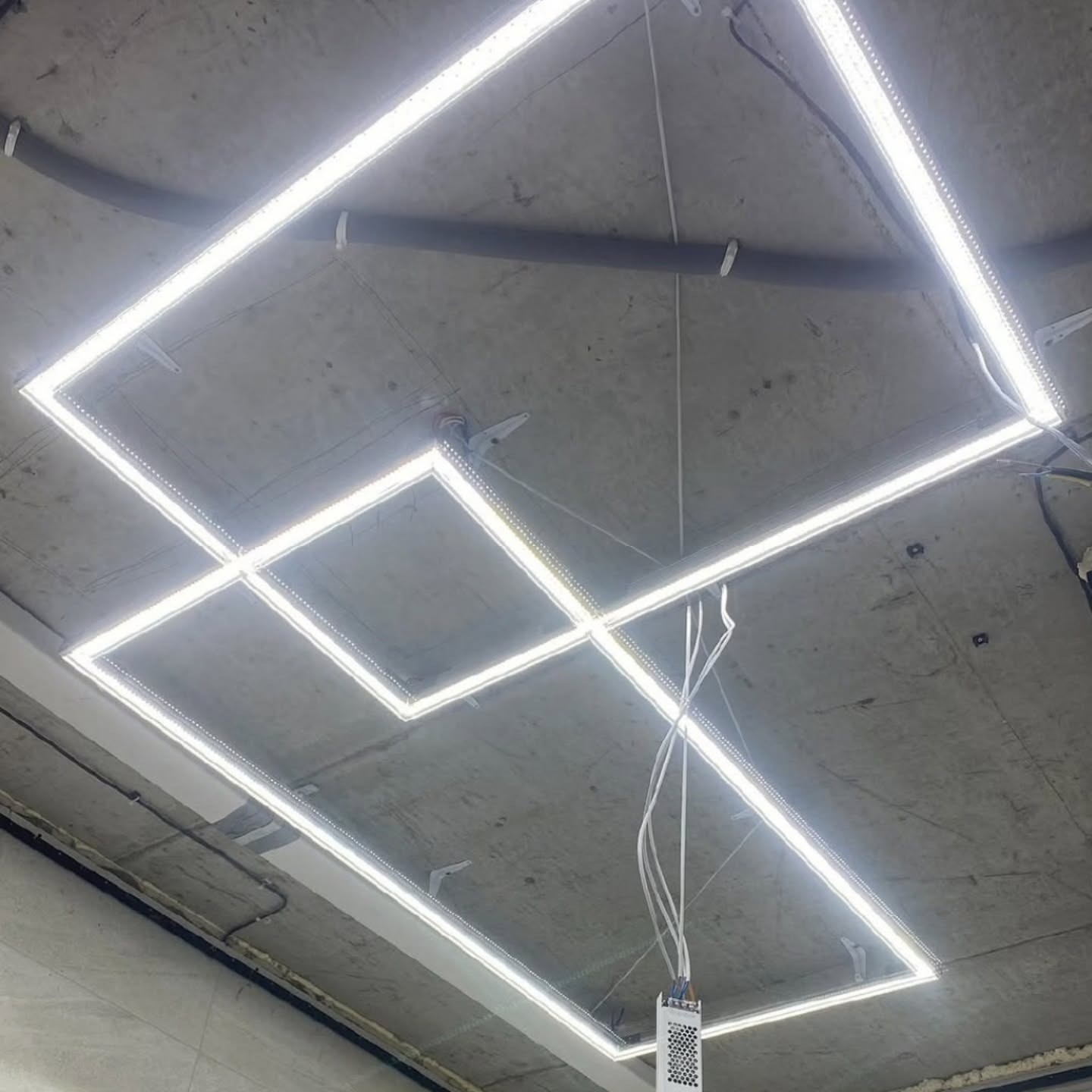 Kwadratowy układ paneli LED zamontowany na surowym, betonowym suficie. Widoczne przewody i elementy mocujące. Oświetlenie równomierne, jasne.