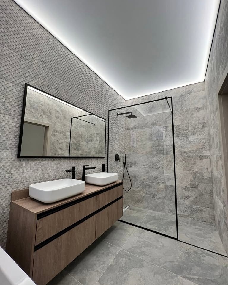 Nowoczesna łazienka z szarą mozaiką, drewnianą szafką pod umywalki i kabiną prysznicową typu walk-in z czarnymi ramami, oświetlona taśmą LED.