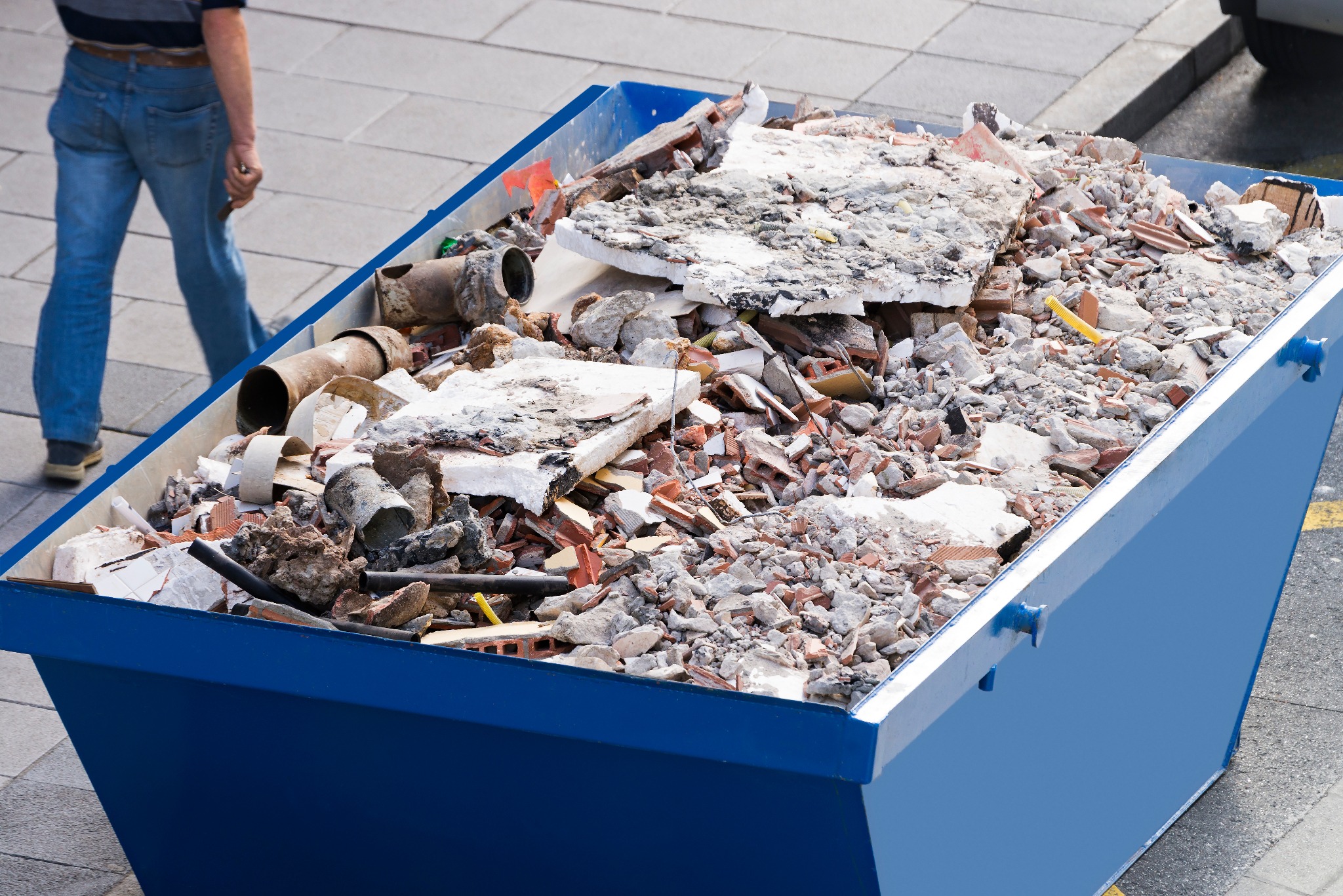 Niebieski kontener wypełniony gruzem budowlanym, w tle fragment chodnika i osoba. Widoczne elementy instalacji, cegły i fragmenty betonu.