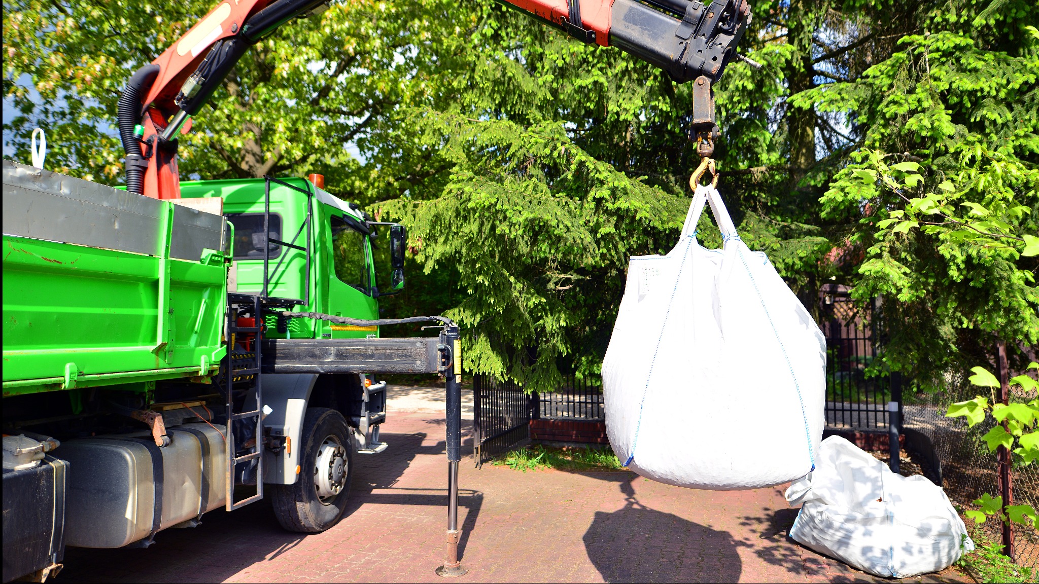 Zielona ciężarówka z dźwigiem podnosi biały worek typu Big Bag na tle zieleni. Widoczny drugi, mniejszy worek obok, na czerwonej kostce brukowej.