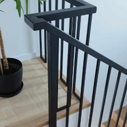 Błażej Owczarek - Jasne, drewniane schody z czarną, nowoczesną balustradą z pionowymi szczeblami. U dołu widoczna roślina doniczkowa. Minimalistyczny design.