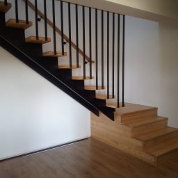 Błażej Owczarek - Klatka schodowa z drewnianymi stopniami i czarną, metalową konstrukcją nośną oraz balustradą z pionowych, czarnych prętów. Jasne drewno stopni kontrastuje z bielą ścian.