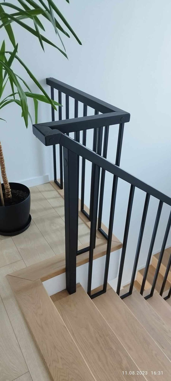 Jasne, drewniane schody z czarną, nowoczesną balustradą z pionowymi szczeblami. U dołu widoczna roślina doniczkowa. Minimalistyczny design.