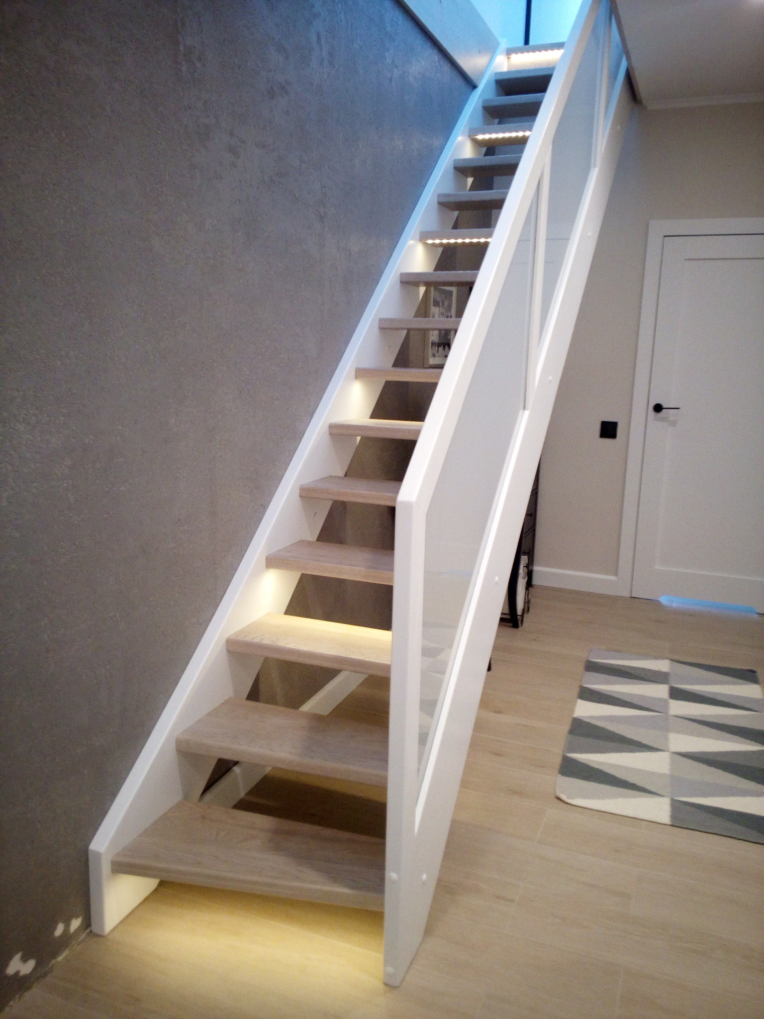 Jasne, minimalistyczne schody z drewnianymi stopniami i białymi balustradami, podświetlone od spodu, prowadzące do góry. Obok szara ściana i białe drzwi.