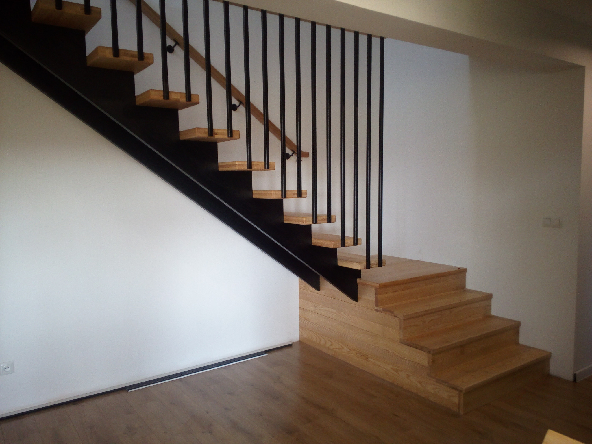 Klatka schodowa z drewnianymi stopniami i czarną, metalową konstrukcją nośną oraz balustradą z pionowych, czarnych prętów. Jasne drewno stopni kontrastuje z bielą ścian.