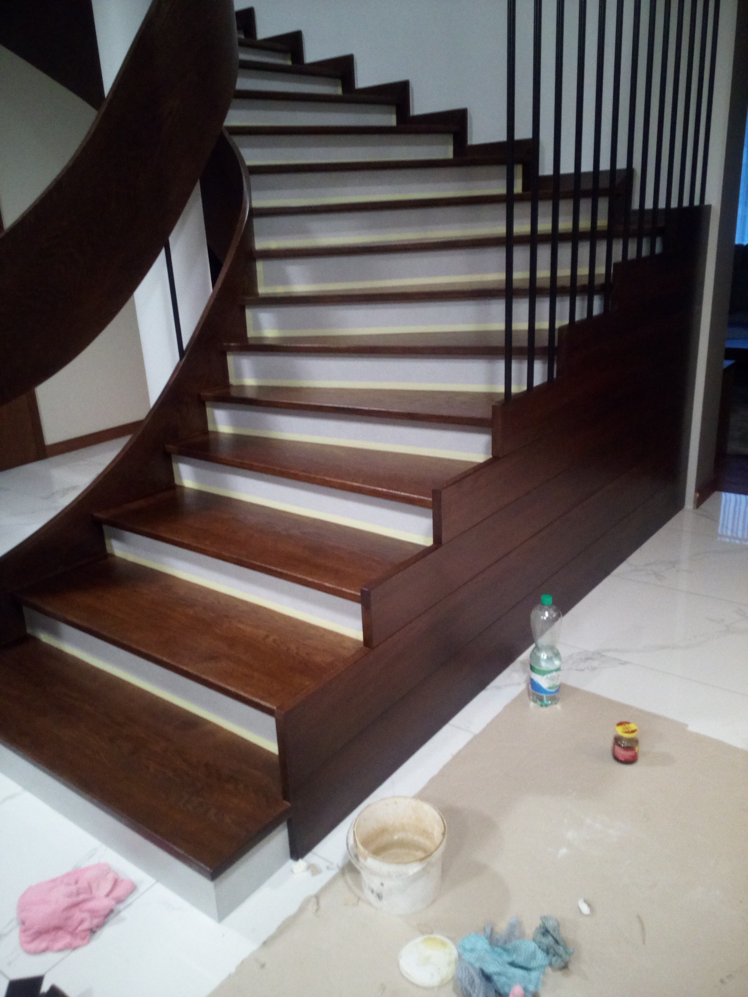 Wykończone drewniane schody z czarną, metalową balustradą. Widoczne narzędzia malarskie i zabezpieczenia na stopniach. Podłoga wyłożona białymi płytkami.