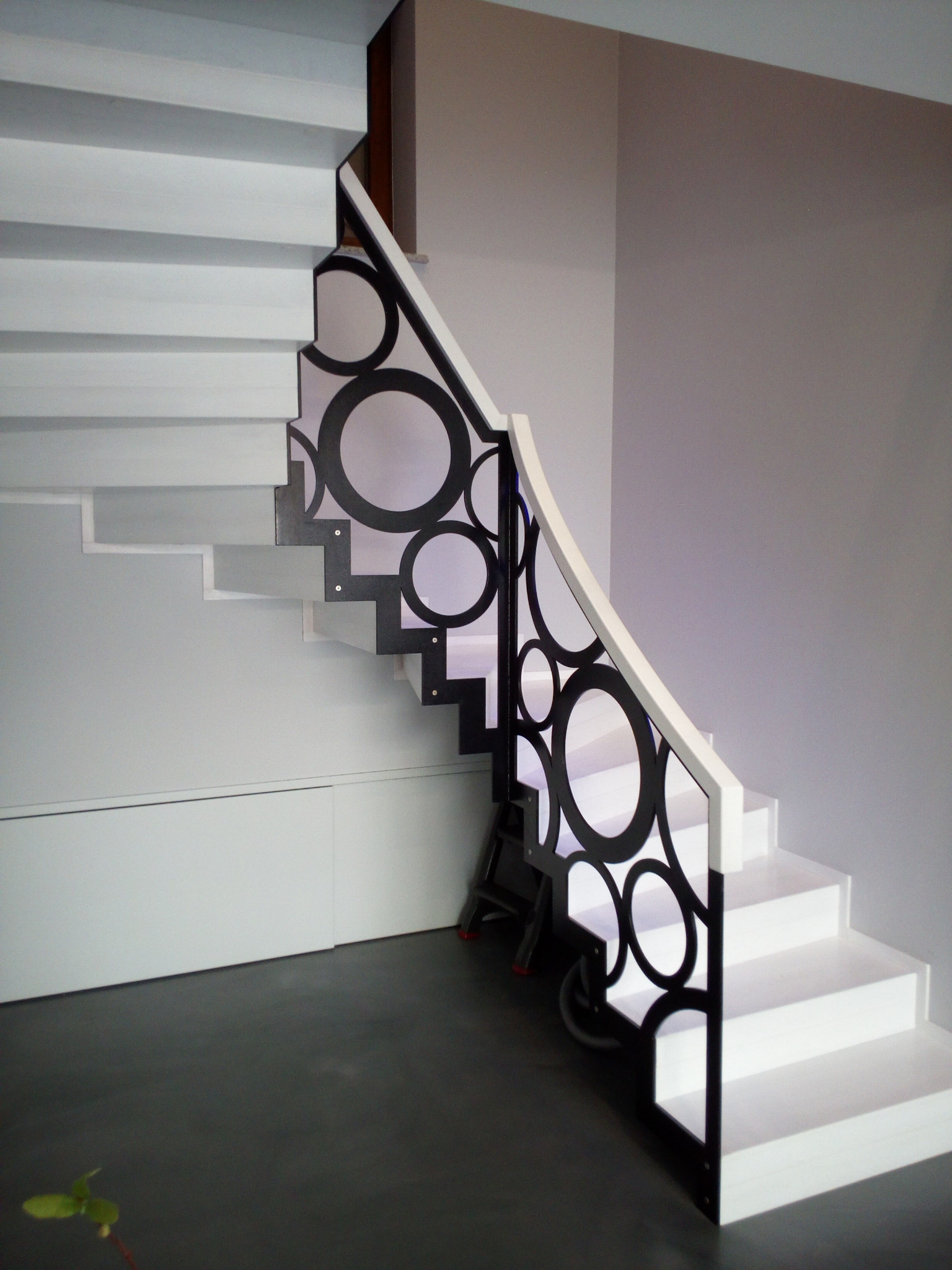 Białe schody z czarną, ażurową balustradą o wzorze z okręgów. Nowoczesny design, subtelne oświetlenie, minimalistyczne wnętrze. Widoczny fragment drabiny.