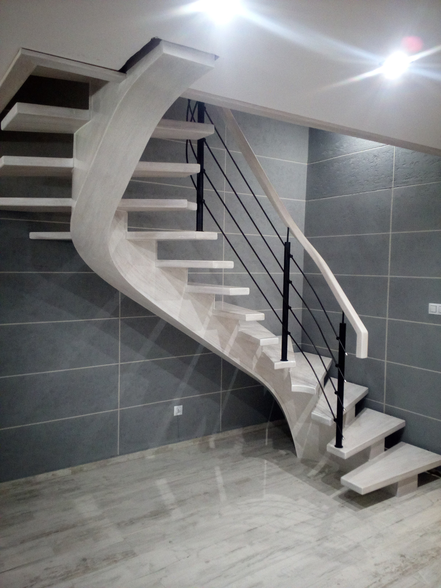 Kaskadowe schody drewniane o nietypowym, zakrzywionym kształcie z czarnymi metalowymi balustradami na tle szarej ściany z płytek. Nowoczesny design.