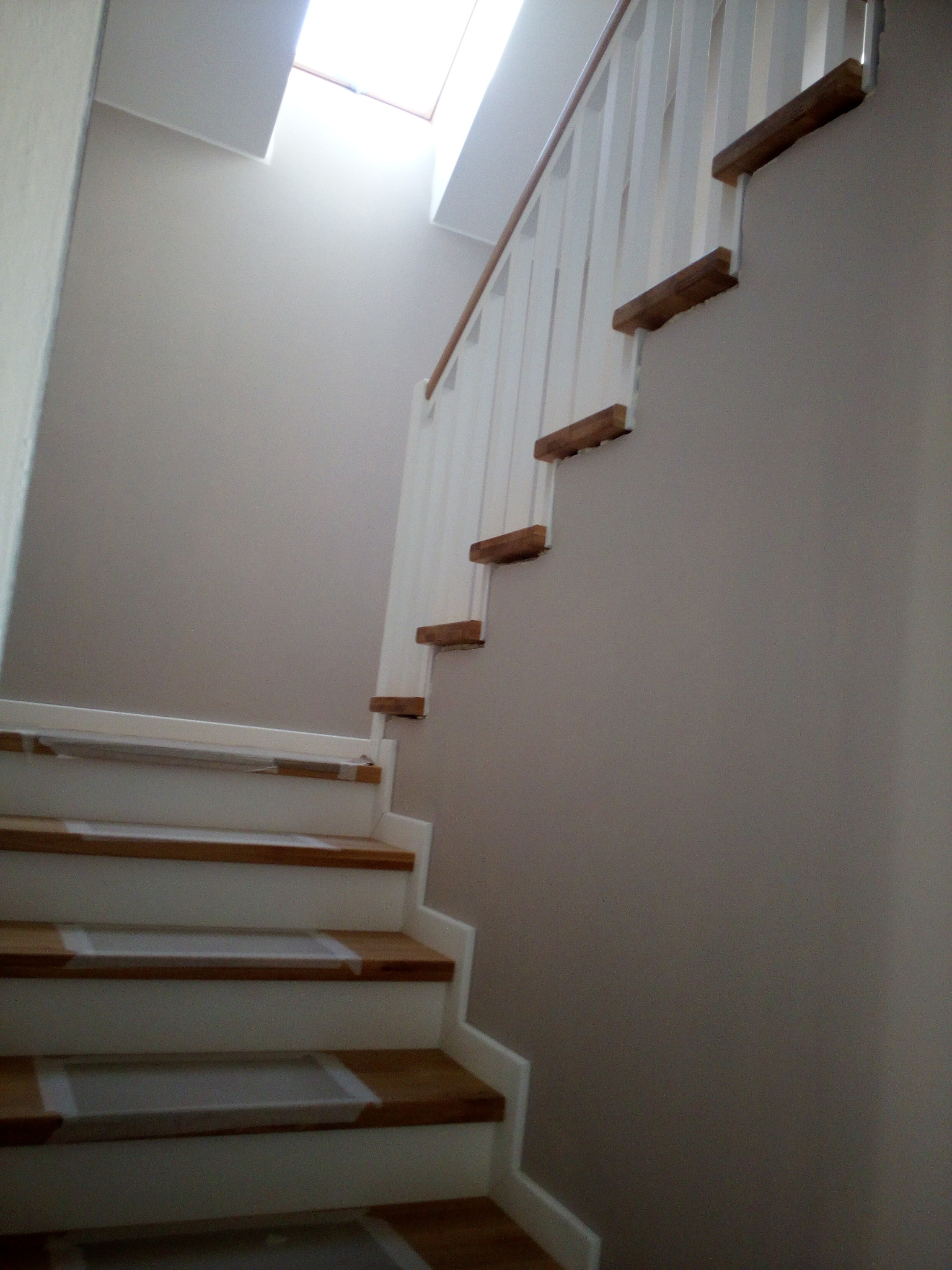 Białe schody z drewnianymi stopniami i balustradą, oświetlone przez okno dachowe. Widok z dołu, z bliska na dolne stopnie zabezpieczone taśmą malarską.