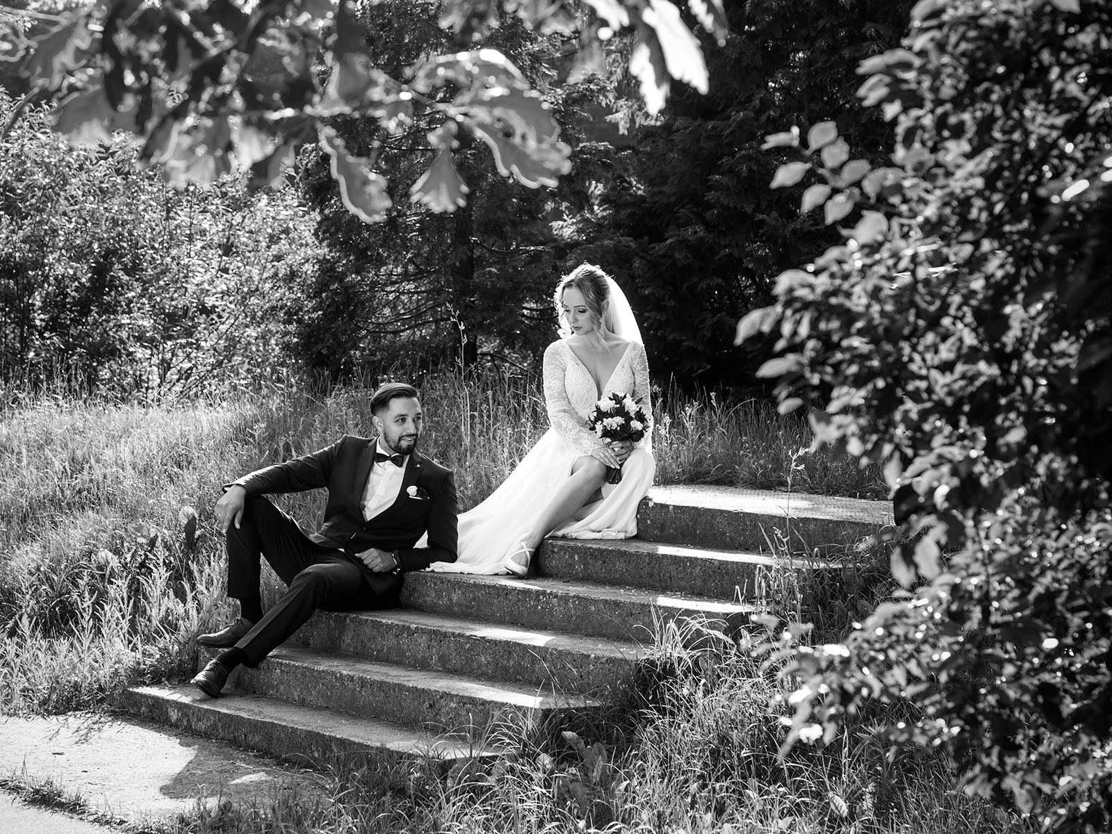 Czarno-białe zdjęcie pary młodej siedzącej na betonowych schodach w plenerze. Pan młody w garniturze, panna młoda w sukni ślubnej z bukietem. Liście drzew w tle i na pierwszym planie.