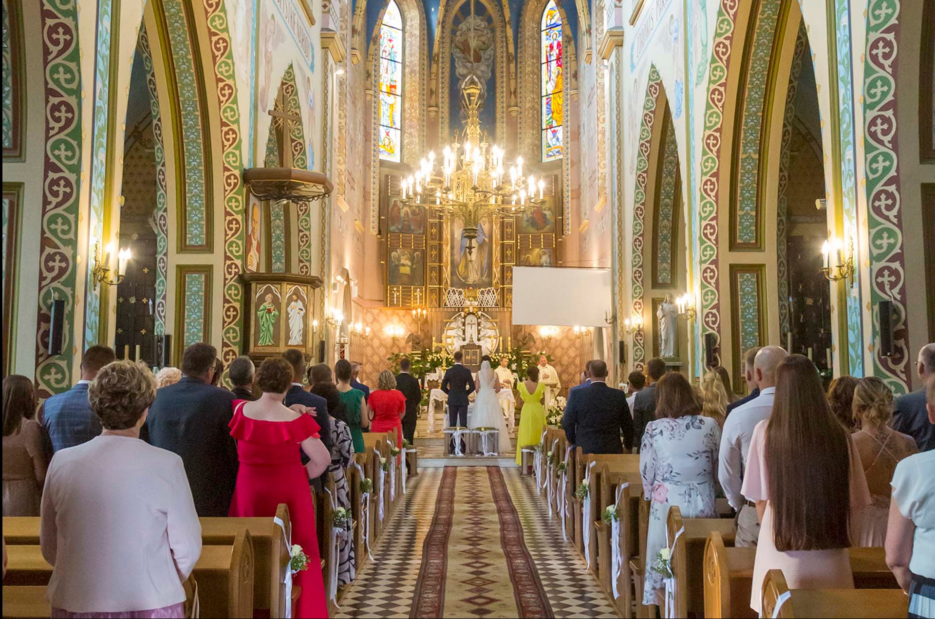 Para młoda podczas ceremonii ślubnej w bogato zdobionym kościele z wysokimi łukami, witrażami i żyrandolem. Goście zgromadzeni w ławkach.
