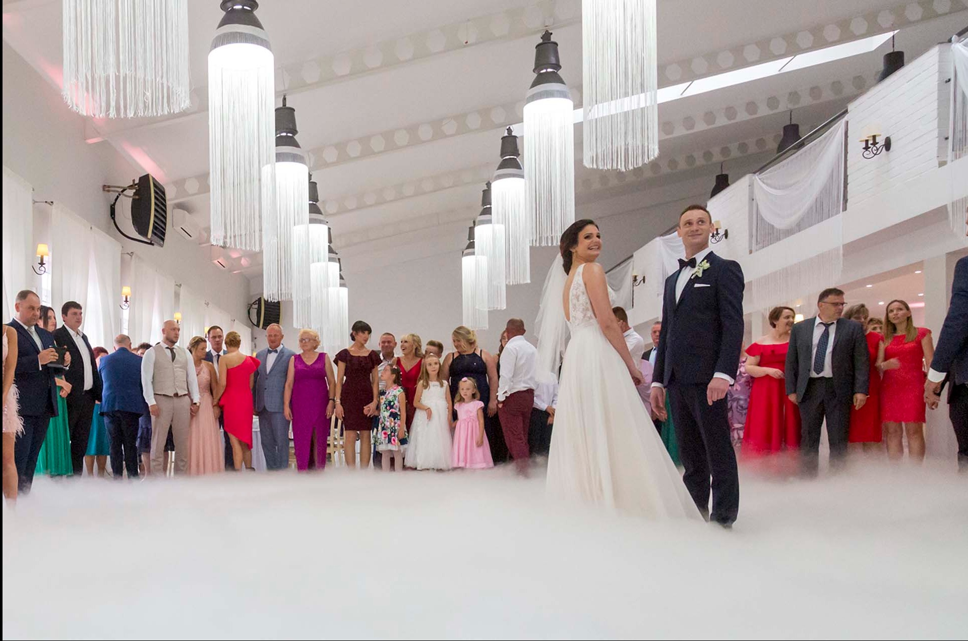 Para młoda w otoczeniu gości weselnych, z efektem ciężkiego dymu na parkiecie. W tle dekoracje sali z wiszącymi lampami z frędzlami i białymi ścianami.