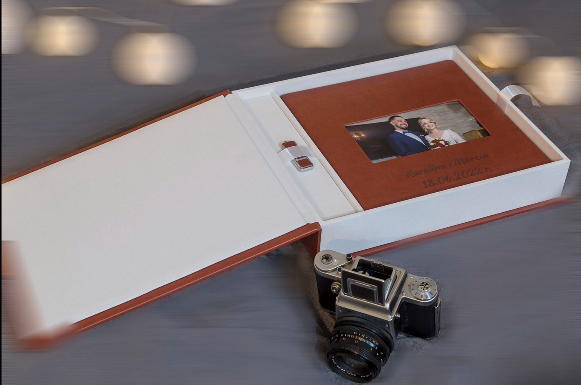 Otwarty, elegancki album ślubny w pudełku z wygrawerowanymi imionami i datą, obok leży klasyczny aparat fotograficzny. Tło rozmyte, z efektem bokeh.