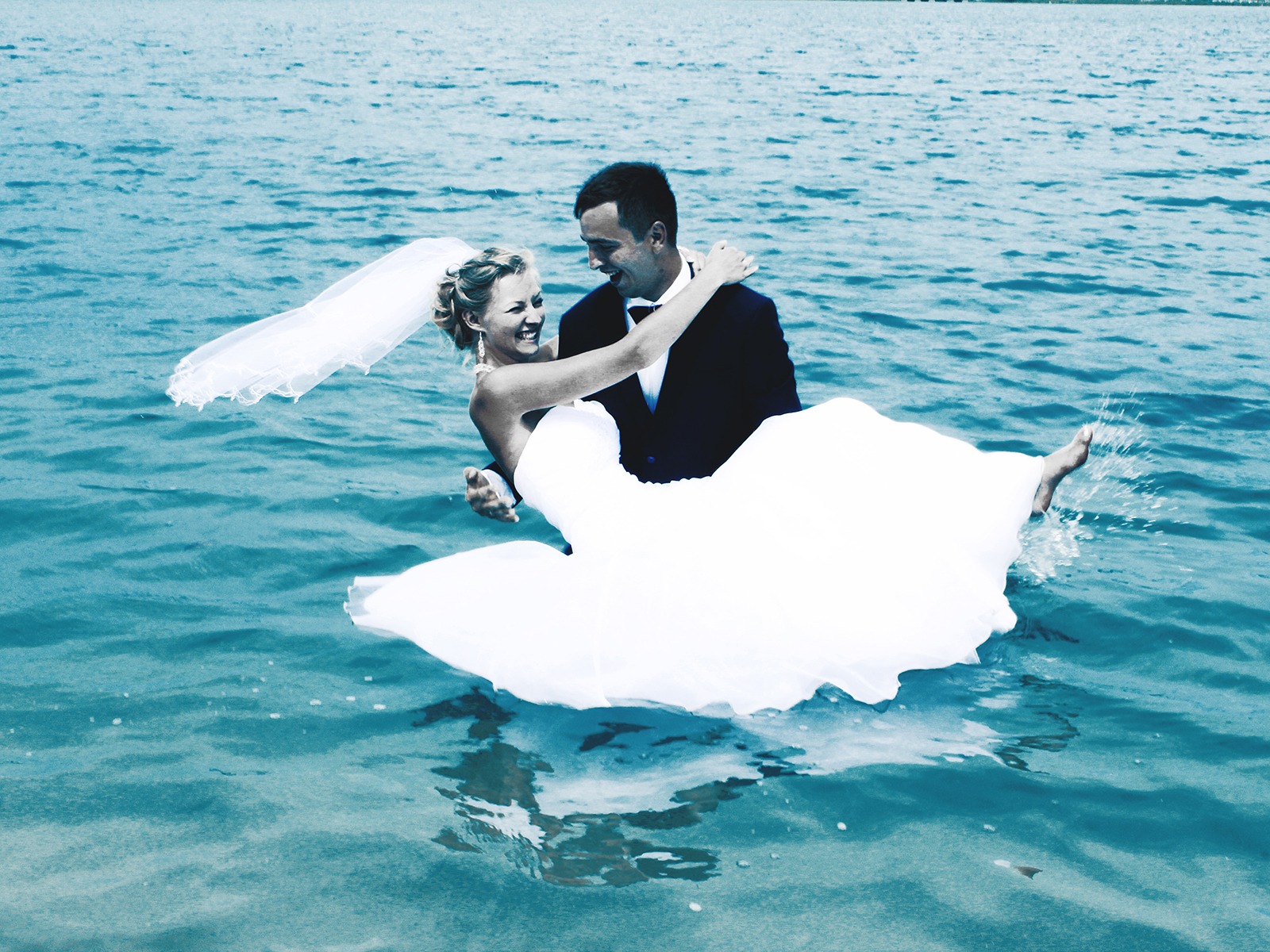 Uśmiechnięta para młoda w wodzie: panna młoda w objęciach pana młodego, woda pryska spod stopy. Nietypowa sesja ślubna w plenerze, jasne, chłodne barwy.