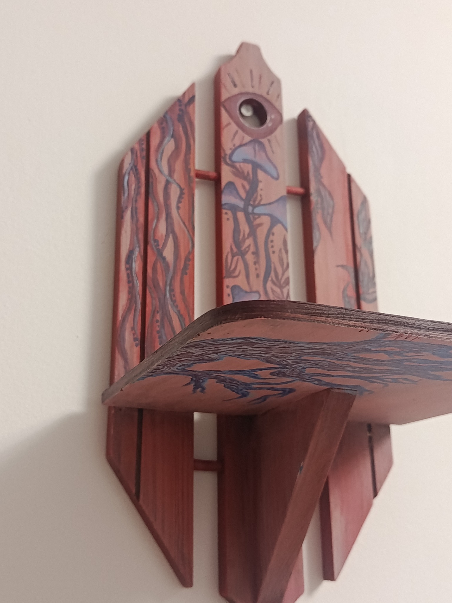 Ręcznie malowana, drewniana półka w stylu boho z motywami roślinnymi i okiem, zawieszona na białej ścianie. Unikalny design i artystyczne wykończenie.