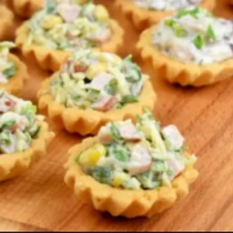 Kruche babeczki z sałatką jarzynową na desce. Widoczne kukurydza, groszek i szynka. Apetyczne przekąski na przyjęcie, idealne na catering firmowy lub świąteczny.