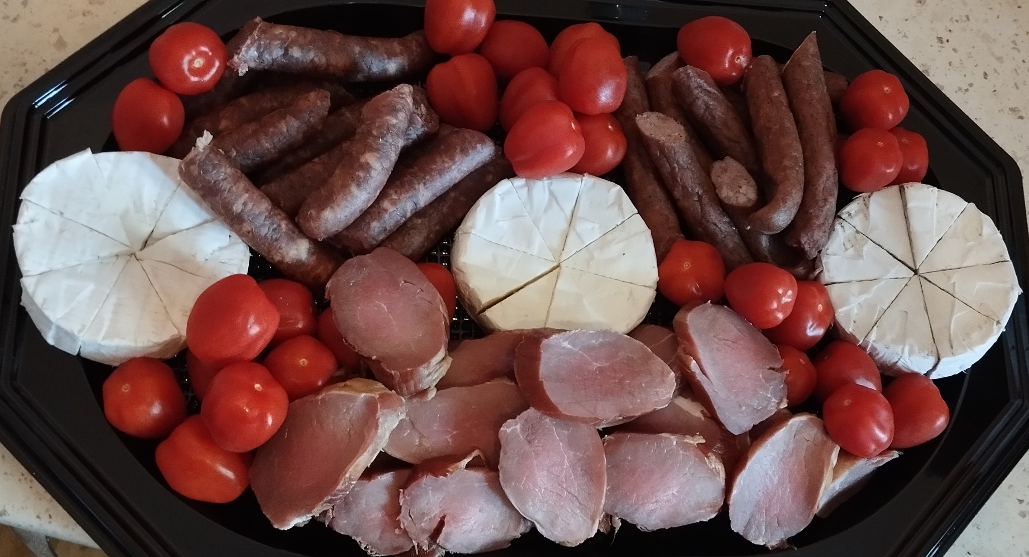 Dekoracyjna, czarna taca z serami brie (jeden pokrojony), wędlinami, plastrami pieczonego mięsa i świeżymi pomidorkami koktajlowymi. Idealna przekąska na imprezy.