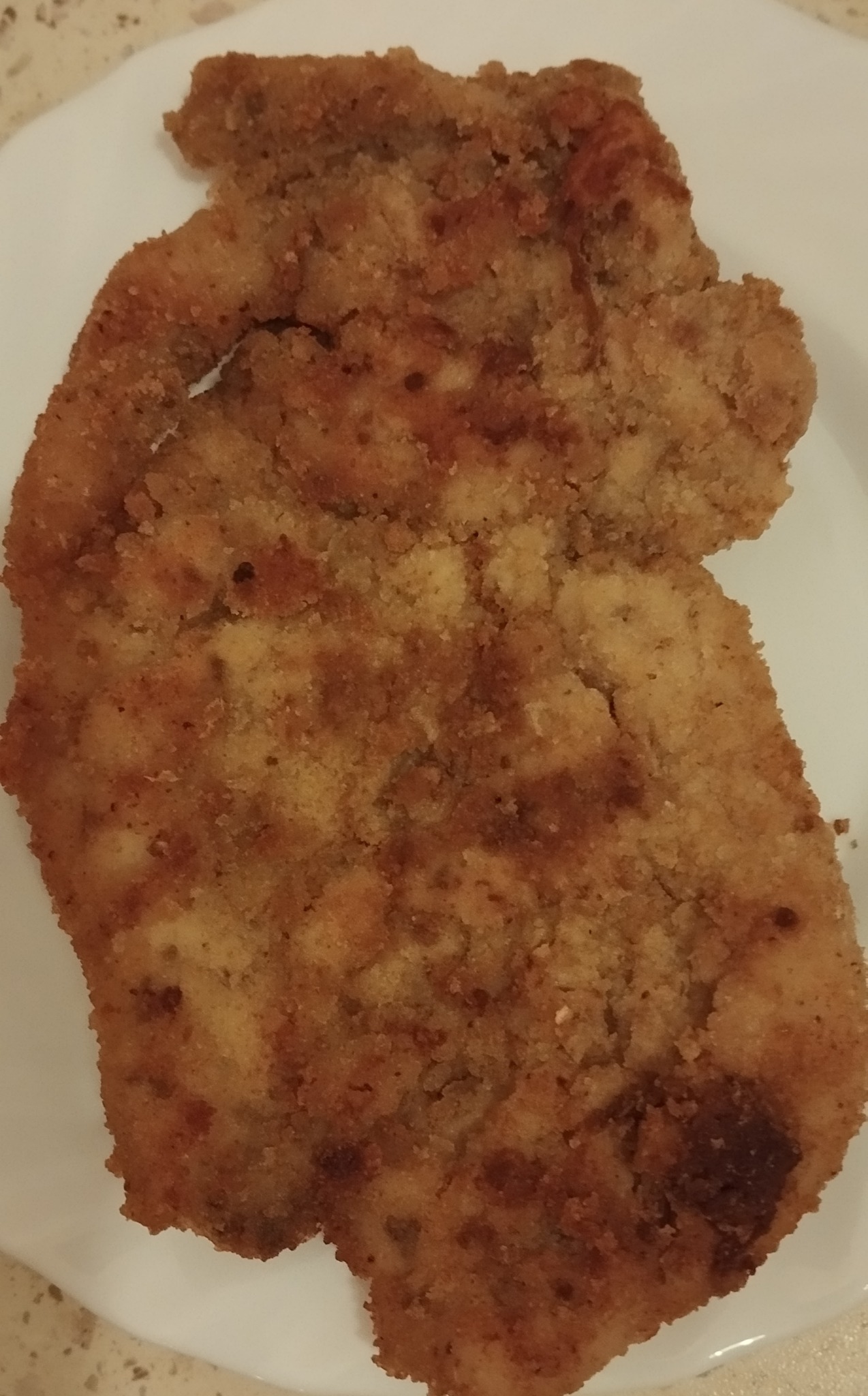 Apetyczny, złocisty sznycel w panierce na białym talerzu, gotowy do spożycia. Widoczne nierównomierne zarumienienie panierki i chrupiąca tekstura.