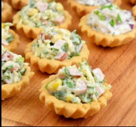 Kruche babeczki z sałatką jarzynową na desce. Widoczne kukurydza, groszek i szynka. Apetyczne przekąski na przyjęcie, idealne na catering firmowy lub świąteczny.