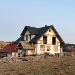 Dom w stanie surowym otwartym z czarnym dachem na tle pagórkowatego krajobrazu. Widoczne drewniane elementy konstrukcyjne i prowizoryczne zabezpieczenia okien.