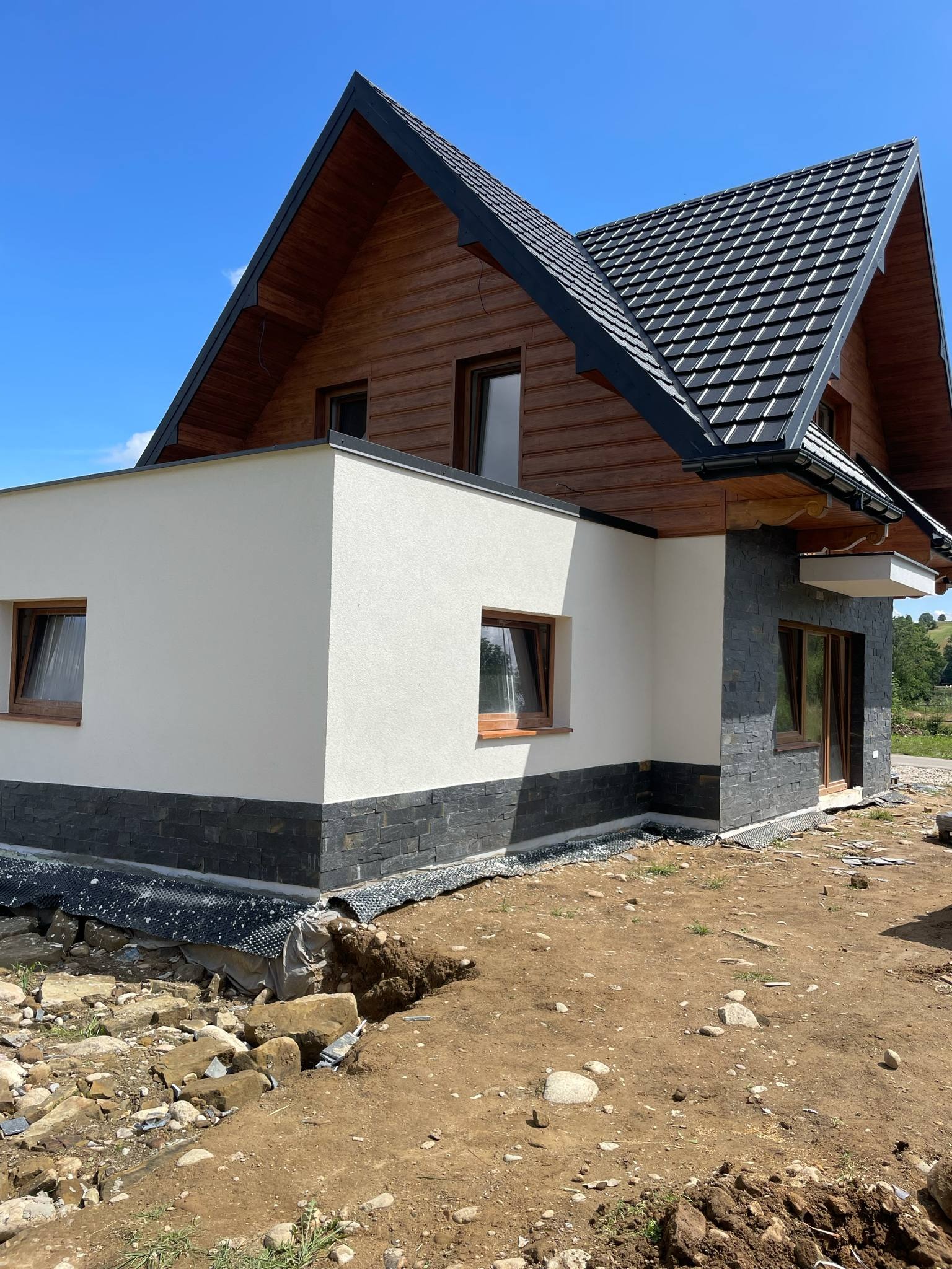 Nowoczesny dom z drewnianą elewacją i czarnym dachem, wykończony kamieniem dekoracyjnym. Widoczne okna z drewnianą stolarką i fragment terenu wokół domu.