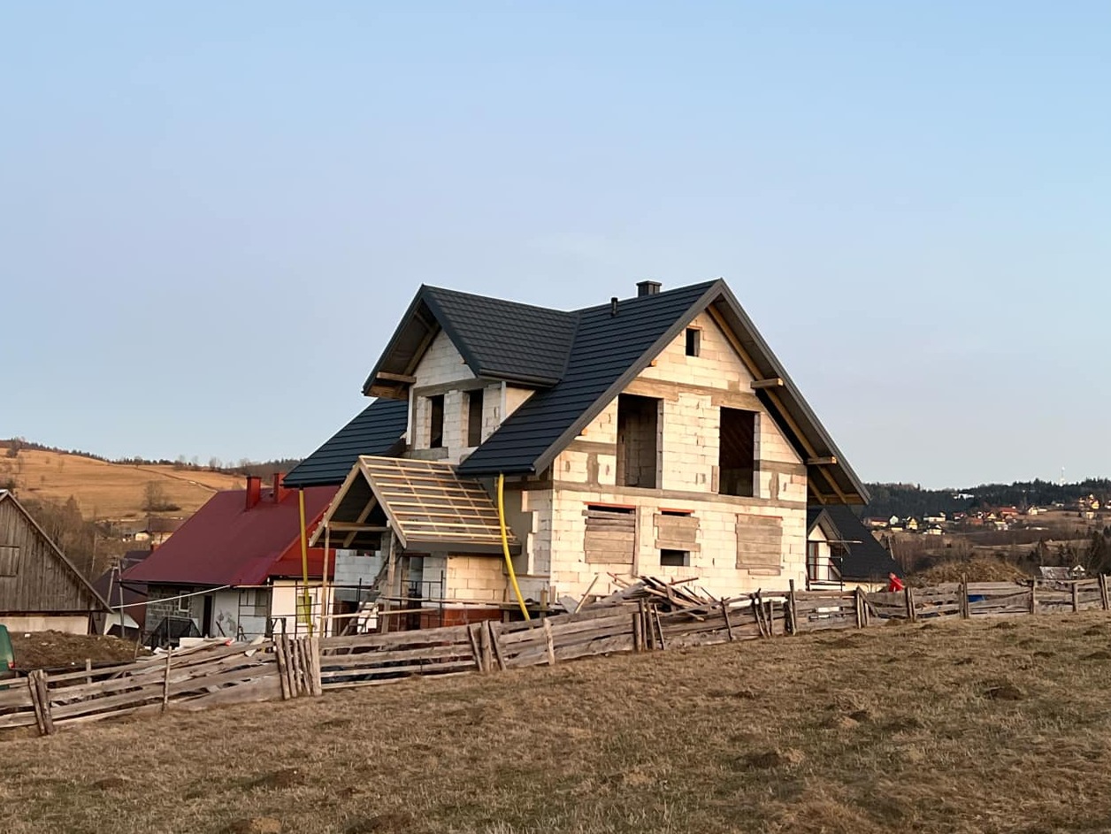 Dom w stanie surowym otwartym z czarnym dachem na tle pagórkowatego krajobrazu. Widoczne drewniane elementy konstrukcyjne i prowizoryczne zabezpieczenia okien.
