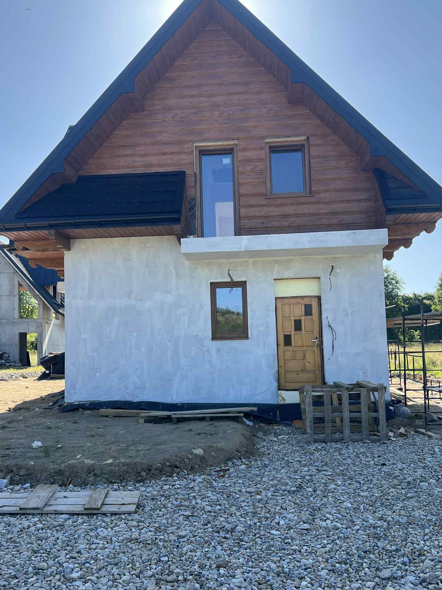 Surowy stan domu z drewnianą górą i otynkowaną dolną częścią. Widoczne okna i drzwi wejściowe. Teren wokół domu wyłożony kamieniami.