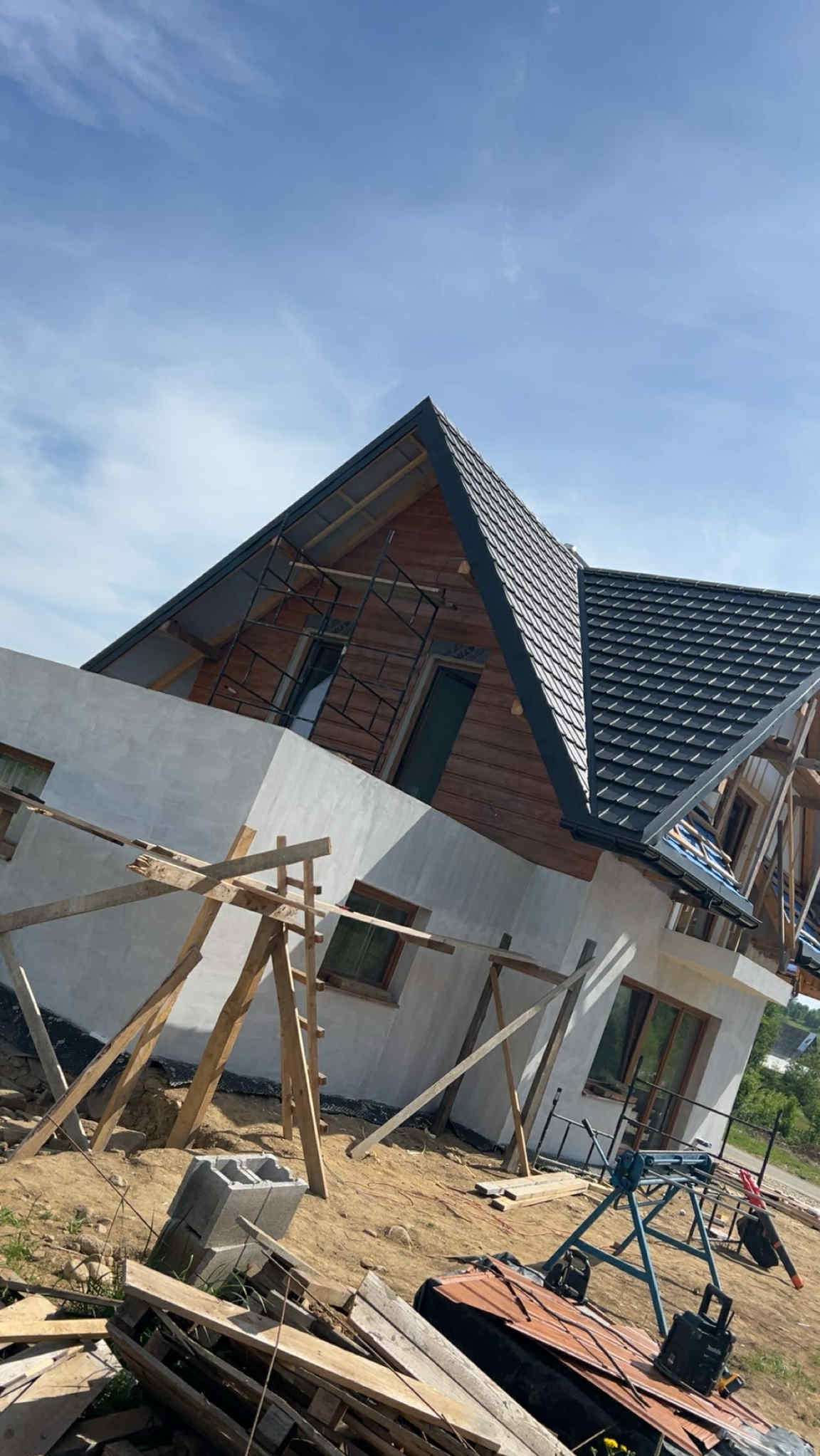 Budowa domu z drewnianymi elementami i szarą dachówką, widoczne rusztowania i materiały budowlane. Częściowo otynkowana elewacja, w tle błękitne niebo.