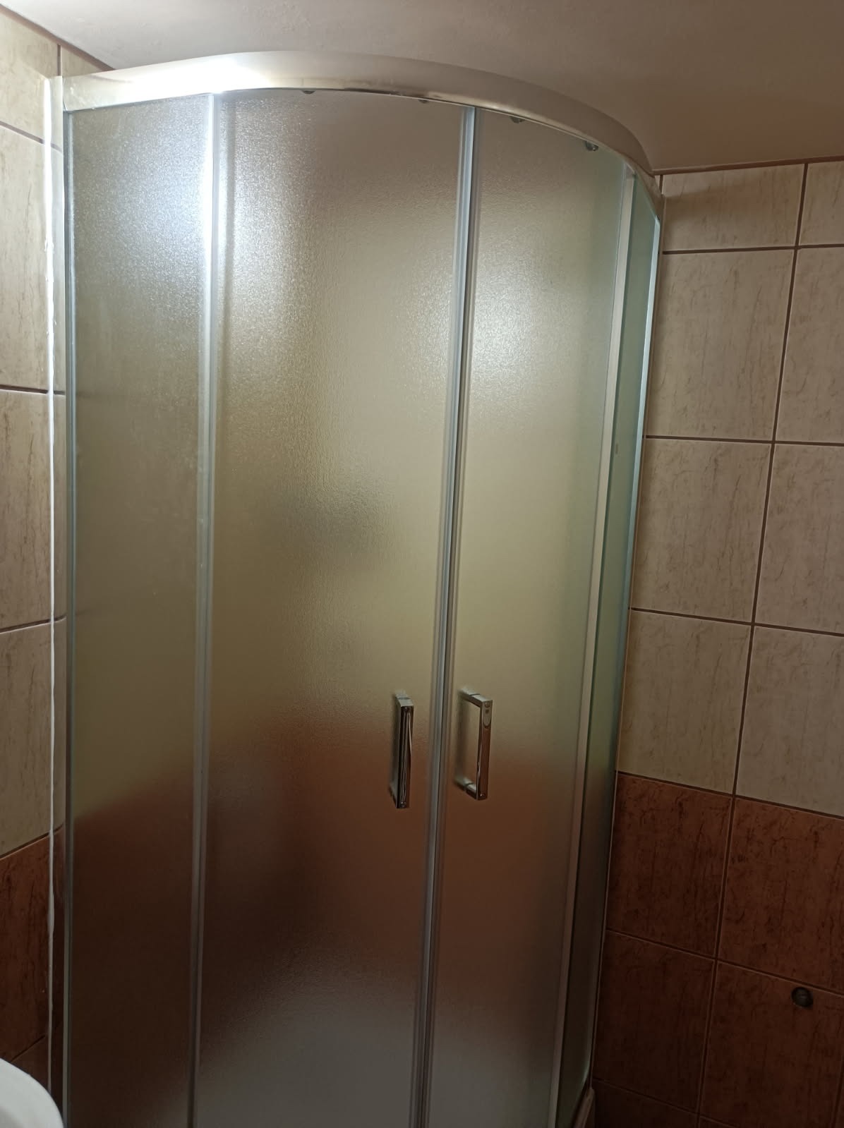 Kabina prysznicowa z matowego szkła w narożniku łazienki, widoczne chromowane uchwyty i płytki w odcieniach beżu i brązu. Widok na zamontowaną kabinę.