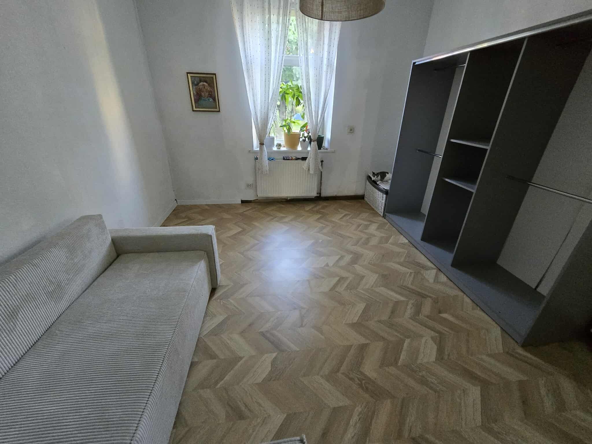 Salon po remoncie: parkiet w jodełkę, szara sofa, szafa wnękowa, okno z roślinami. Widok na jasne, odnowione wnętrze z elementami dekoracyjnymi.