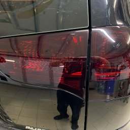BLACK STUDIO - Ciemnoczerwone, przyciemnione tylne światło czarnego samochodu. Widoczne odbicie osoby wykonującej usługę.